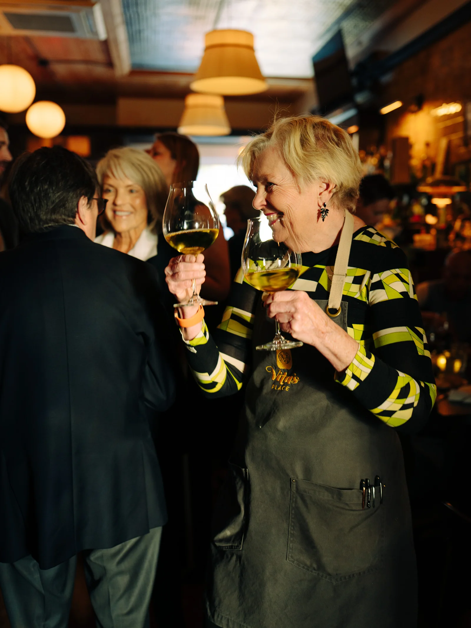A smiling host carrying wine glasses through a lively bar